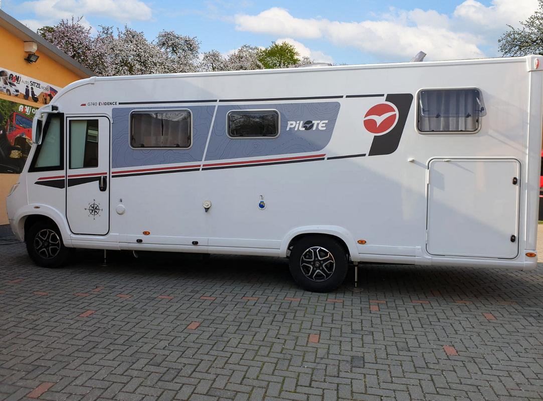 White motorhome with blue graphics and a red Pilote logo parked on a brick lot, side view showing windows and storage compartments